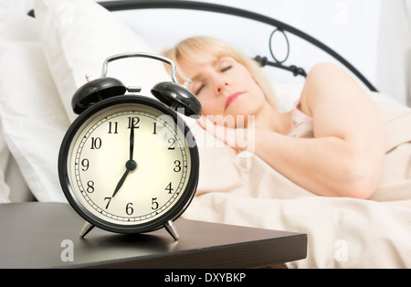 Eine Frau mittleren Alters schläft, während Wecker im Vordergrund 07:00 liest. Stockfoto