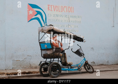 Ein Fahrrad-Taxi-Fahrer eine Pause durch revolutionäre Propaganda in zentralen Havanna, Kuba. Stockfoto