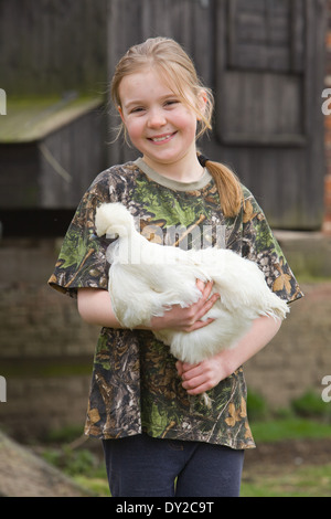 Ein junges Mädchen hält ein weißes Huhn Silkie außerhalb mit einem Holzhaus Henne im Hintergrund Stockfoto