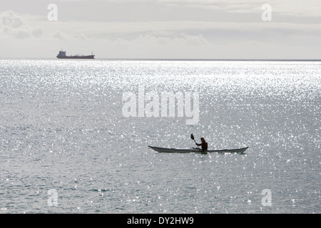 Ein Kajakfahrer sitzt in seinem Kajak Maenporth Strand, Falmouth, während ein Schiff am Horizont sitzt. Stockfoto