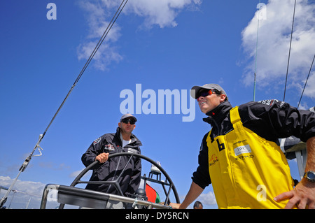 Kapitän am Steuer eines Ozeans Rennyacht mit einem Crew-Mann stehend in der Nähe, tragen Sonnenbrillen und Kappen unter blauem Himmel Stockfoto