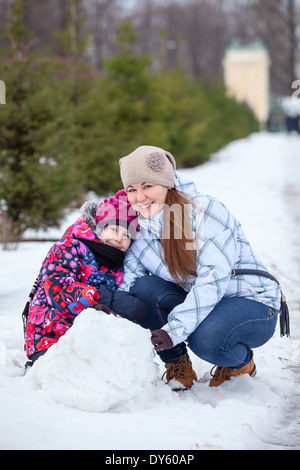 Glückliche Mutter mit Tochter sitzen im Schnee mit Schneeball im Winter park Stockfoto