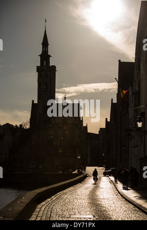 BRÜGGE, Belgien – Ein einsamer Radfahrer durchquert eine historische Kopfsteinpflasterstraße entlang eines der malerischen Kanäle Brügge. Die Silhouette der Figur vor dem mittelalterlichen Stadtbild spiegelt den zeitlosen Charakter der UNESCO-Weltkulturerbe-Stadt wider. Diese atmosphärische Szene veranschaulicht die Erhaltung historischer Straßenmuster und traditioneller Transportmethoden im modernen Brügge. Stockfoto