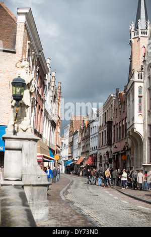 Wollestraat mittelalterliche Straße Brügge Belgien | BRÜGGE, Belgien – Fußgänger und Radfahrer bewegen sich entlang der Wollestraat, einer kopfsteingepflasterten Durchgangsstraße, die den Markt mit der Burg unter aufstrebenden Sturmwolken verbindet. Die Stufengiebel-Ziegelfassaden der Straße spiegeln den neo-Brügge-Restaurierungsstil wider, der weit über das historische Zentrum verbreitet wurde. Eine Statue auf einer Lampenpfosten-Halterung umrahmt den Blick auf den mittelalterlichen Stadtkern. Stockfoto