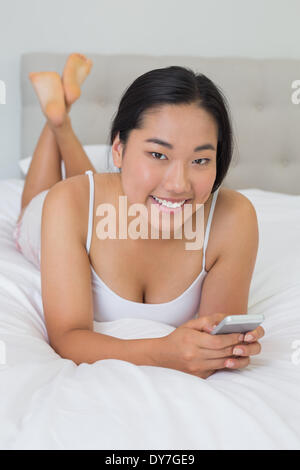 Lächelnde asiatische Frau liegend auf Bett SMS auf Handy Stockfoto