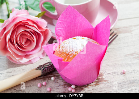 Muffin in rosa Wickel, Rosenblüten, Tasse, Gabel und Zucker Perlen auf Holztisch Stockfoto