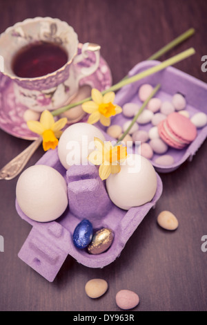 Gedeckten Tisch mit Tasse Tee, egg Narzissen (Narcissus Pseudonarcissus), Carton Macaron, drei Eiern und Süßigkeiten auf Holztisch Stockfoto