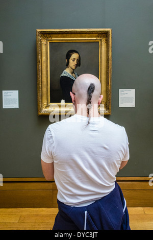 Ein junges Männchen mit einem rasierten Kopf und ein Pony tail Haar Ansichten ein Gemälde an der Wand eines Museums. Stockfoto