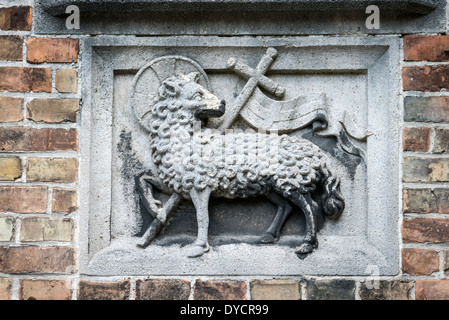 BRÜGGE, Belgien — Eine geschnitzte Tafel mit dem Agnus Dei (Lamm Gottes), Symbol des heiligen Johannes des Täufers, ziert das Äußere des Sint-Janshospitaal (altes Johanniskrankenhaus). Dieses religiöse Emblem, kombiniert mit einem Kreuz, kennzeichnet die Widmung des Gebäudes an seinen schutzheiligen. Das architektonische Detail repräsentiert die doppelte Rolle des Krankenhauses sowohl als medizinische als auch als religiöse Institution. Stockfoto