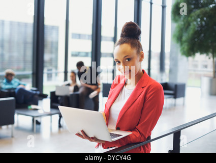 Schöne junge Frau mit einem Laptop im Büro. Afrikanische amerikanische Frau, die durch ein Geländer mit Kollegen im Hintergrund. Stockfoto