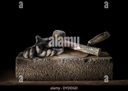 Des Erbauers Ziegel Kelle mit Lump Hammer und Handschuhen ruht auf einem Betonblock. Stockfoto