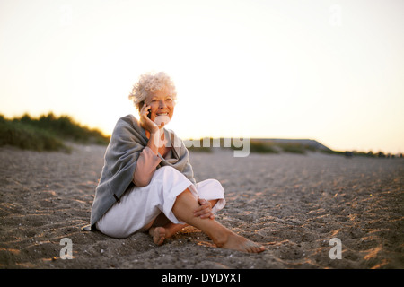 Entspannt im Ruhestand Trägerin Schal sitzt am Sandstrand einen Anruf tätigen. Alten kaukasischen Frau sitzt auf der Strand suchen Stockfoto