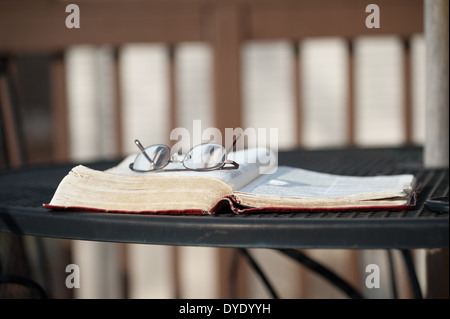 Ein paar der Lesebrille liegt oben auf eine aufgeschlagene Bibel Verlegung auf einem schmiedeeisernen Tisch auf einem Holzdeck mit passenden Stühlen. Stockfoto