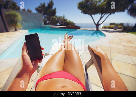 Junge Dame mit Bikini mit Handy beim Sonnenbaden am Pool. Weibliches Modell ruht auf einem Liegestuhl mit Smartphone Stockfoto