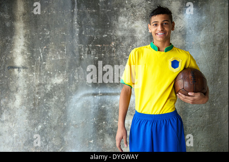 Lächelnde junge brasilianische Fußballspieler in Team-Uniform von Brasilien Farben halten Vintage Fußball gegen konkrete Favela Wand Stockfoto