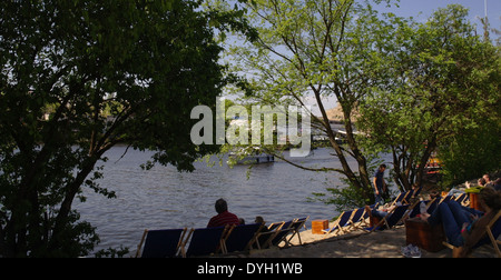 Menschen, die Liegestühle auf Sand unter grünen Bäumen sitzen mit Blick auf Fluss Spree, Strandbar Oststrand, East Side Gallery, Berlin Stockfoto