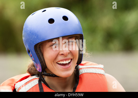 Eine Nahaufnahme einer Frau mit blauem Schutzhelm mit Belüftungslöchern und orangefarbener Schwimmweste mit reflektierenden Streifen. Sie lächelt breit, zeigt ihre Zähne, mit geknitterten Augen Stockfoto