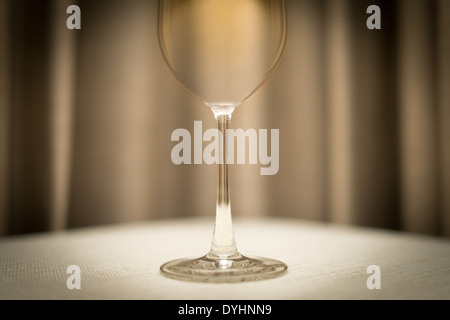 Nahaufnahme der leere Weinglas auf Tisch mit weißer Tischdecke. Glänzende einziges Glas. Verschwommene braunen Vorhang im Hintergrund. Stockfoto