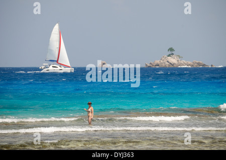 Segelboot angesehen vom schweren Strand Anse mit Frau am Riff im Vordergrund auf La Digue, Seychellen Stockfoto