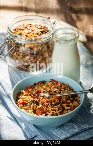 Portion hausgemachtem Müsli in blaue Schale und Milch oder Joghurt auf Tisch mit Bettwäsche Stockfoto