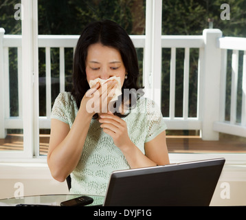 Foto von Reife Frau ihre Nase mit Gewebe während der Arbeit zu Hause mit Laptop und Handy auf Tisch abwischen Stockfoto