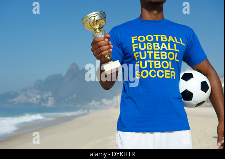 Champion brasilianischer Fußballspieler mit Trophäe und Soccer Ball im internationalen Fußball T-shirt Ipanema Strand Rio De Janeiro Stockfoto