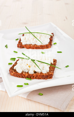 Schwarzes Vollkornbrot Scheiben mit Frischkäse, frischen Kräutern und Weizen auf weißen Teller. Gesundes Essen rustikal Stockfoto