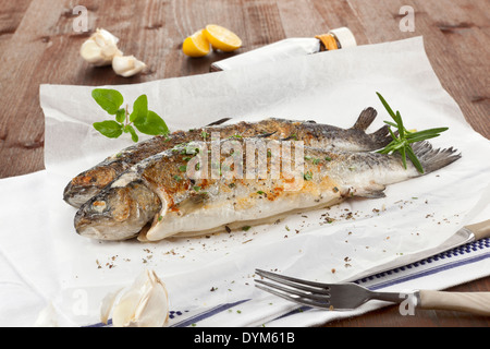 Zwei gegrillte Forelle mit Zitrone, frischen Kräutern und Knoblauch auf Backpapier auf Küchentuch auf Holztisch weiß. Stockfoto