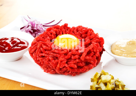 Steak Tartar mit Eigelb auf weißen Teller mit Zwiebeln, Gurken, Senf und Ketchup. Toast im Hintergrund. Stockfoto