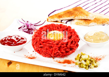 Steak Tartar mit Eigelb auf weißen Teller mit Zwiebeln, Gurken, Senf und Ketchup. Toast im Hintergrund. Stockfoto