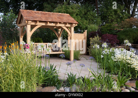 Englischer Cottage Garten mit Eiche gerahmter Laube Holzbank Sitzschwinge Holztor Stein gepflasterte Terrasse gemischte Blume Grenzen Grenze blühend im Sommer Großbritannien Stockfoto