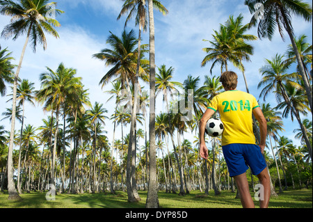 Brasilianischer Fußballspieler in Brasilien Farben einheitliche Stellung im Hain von tropischen Palmen halten Fußball Stockfoto