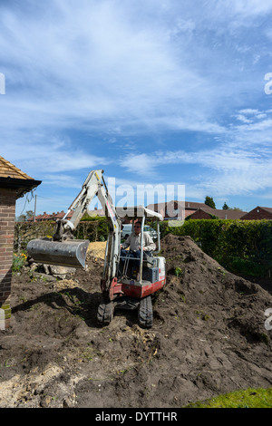 Ein Arbeiter gräbt einen Garten mit einem Mikro Kompaktbagger vor dem Verlegen einer Auffahrt auf eine Renovierung. Weitwinkelaufnahme Stockfoto