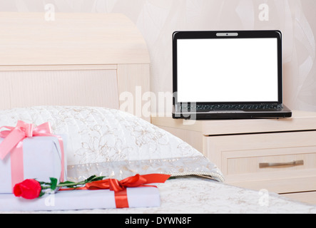 Laptop zu öffnen, mit einem leeren Bildschirm ist auf dem Nachttisch im Schlafzimmer und vor ihm auf dem Bett lag eine Blume und Geschenk-Boxen Stockfoto