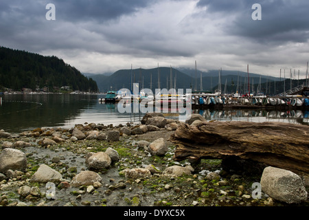 Kieselsteine & Treibholz Vordergrund Steg umgeben mit Boote Yachten & Kanus in noch reflektierende Einlass & hinteren Wald Berge Stockfoto