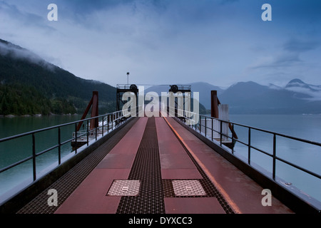 zeigen Sie industrielle Pier Rampe in ruhiges Meer Bucht, umgeben von bewaldeten Bergen, schwebende Nebel deckt meist blauen Himmel BC an Stockfoto