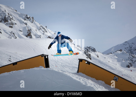 Snowboarder mit wollene Mütze und Brille springt über eine Rampe mit Bergen im Hintergrund. Stockfoto