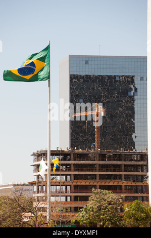 Brasilia, Brasilien. Neubau im Bau mit der brasilianischen Flagge und einem älteren Gebäude mit Glasfront Spiegel abgeschlossen. Arbeiter auf den oberen Etagen und einen Baukran. Stockfoto