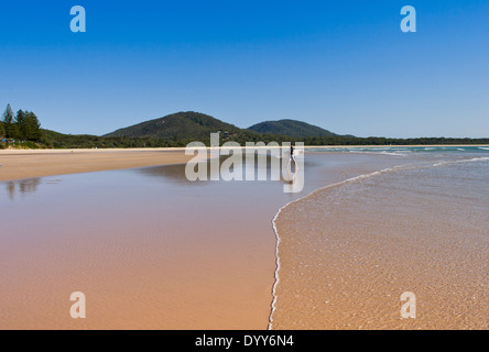 niedrigen Winkel Blick über Wattflächen am Rande Strand mit sanften Welle, Surfer zu Fuß am Strand und weit entfernten Hügel, auf sonnigen Tag Stockfoto