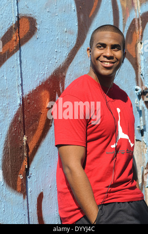 Ein afrikanischer amerikanischer Mann, junger Mann gekleidet im roten T-shirt, Lächeln, glücklich Porträt, Musikhören, Wand gelehnt Stockfoto