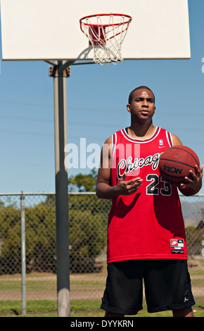 Eine afroamerikanische junger Mann hält einen Basketball vor einen Basketballkorb, stehend auf einem Basketballfeld im Freien. Stockfoto