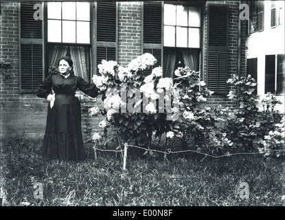 Dieses Foto mit dem Titel „Woman in Keene New Hampshire“ von Bion Whitehouse zeigt eine Frau, die vor einem Backsteinhaus steht, umgeben von blühenden Büschen. Das Bild spiegelt den Kleinstadtcharme von Keene, New Hampshire, wider, mit einem Schwerpunkt auf lokaler Architektur und Natur. Stockfoto