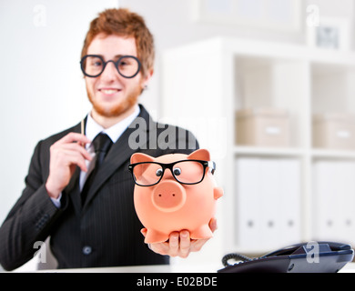 Selbst zuversichtlich lächelnde junge rothaarige Geschäftsmann übergeben Sie ein Sparschwein und beide von Ihnen mit Brille Stockfoto