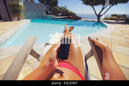 Gebräunte Frau mit Smartphone am Pool. Kaukasische weibliches Modell Sonnenbaden auf einem Liegestuhl und Handy. Stockfoto