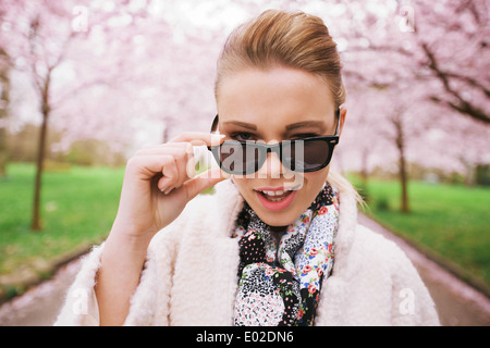 Schließen Sie herauf Bild der netten jungen Frau zwinkert in die Kamera. Freche weibliches Modell im Frühlingspark mit Sonnenbrille. Stockfoto