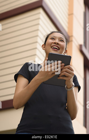 Eine junge asiatische Frau hält eine Ipad, Tablet, Lächeln, wegschauen. Sie trägt ein schwarzes T-shirt. Stockfoto