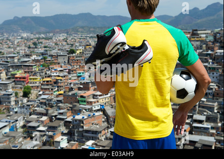 Brasilianischer Fußballspieler stehen in Brasilien-Farben-Kit mit Fußball Ball vor Slum Hintergrund der Favela in Rio De Janeiro Stockfoto