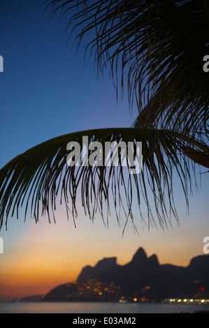 Rio de Janeiro Ipanema Strand Brasilien Sonnenuntergang mit den funkelnden Lichtern der Favela Vidigal und zwei Brüder Berg unter Palme Stockfoto