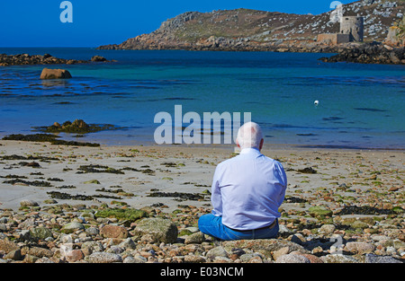 Mann sitzt auf Felsen und Erholung am Strand bei Bryher, Isles of Scilly, Scillies, Cornwall im April mit Cromwells Burg auf Tresco in der Ferne Stockfoto