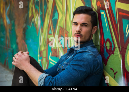 Ernst junger Mann sitzt an bunten Graffiti-Wand, Blick in die Kamera Stockfoto